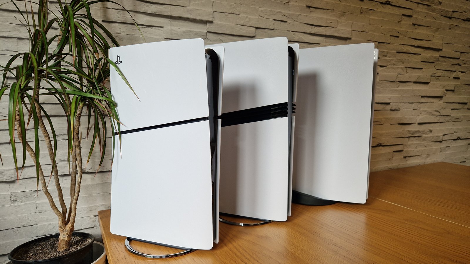 PS5 Slim next to the PS5 Pro and Running PS5 model on a wooden table with white brick and next to a flower vase