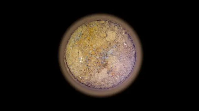 Image of a stone core made on Mars. The picture shows a circular cut of the rock sample on a black background. Orange pattern and has white, gray and black spots.