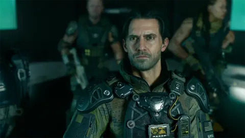 Activision's screenshot shows a realistic rendering of actor Milo Ventimiglia's character, dressed in an elaborate military uniform and body armor. He looks up, his face illuminated by the screen light, his teammates watching in the background.