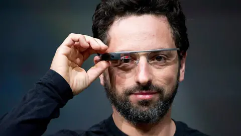 Bloomberg Sergey Brin wears a black long-sleeved T-shirt and Google Glass via Getty Images. He looks straight into the camera lens with one hand resting on the right side of his Google Digital Glasses. On the right lens of the glasses, next to the black box that houses the glasses camera, is a small red and black digital display.