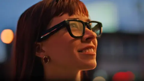 Google close-up of a woman in glasses. She was smiling and had long red hair. The glasses are black with thick frames and the lenses appear to be slightly translucent.